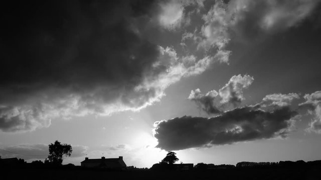 Cumulus in sunset running fast over the countryside. Black and white timelapse footage of cloudscape. Silhouettes of houses, farms, trees in France - Europe.