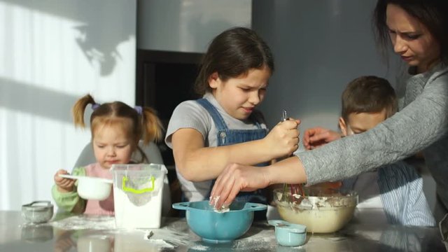 Three children prepare dough in the kitchen. The elder sister helps his brother to stir the dough, the younger plays with flour. Mom dirty the children's noses with flour as a joke. Funny family