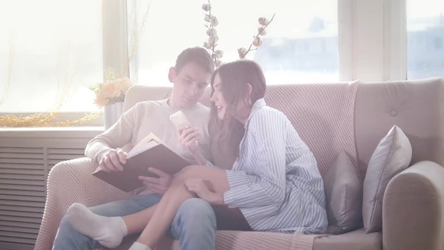 Happy young attractive couple sitting together on the couch, the guy reads a book, the girl use phone