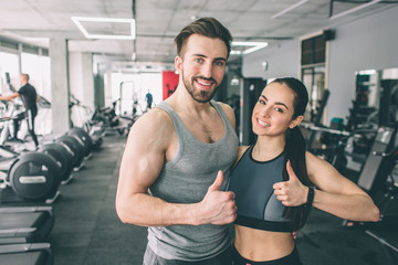 Fototapeta na wymiar Trainer and her student are standing in the sport club's training room together and showing their big thumb's up. They are happy to exsercise in this fitness club. Young couple is smiling.