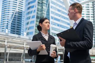 Asian business woman wear suit holding document file and plastic mug on hand talk about business future with  Caucasian businessman in the city.