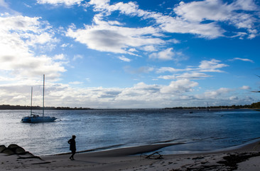 Silhouette of woman walking along the beach at dusk with sailboats and a bridge and trees across the water in the background