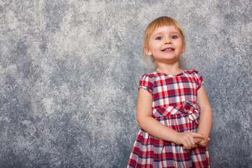 A tiny sweet girl with blond hair wearing a red checkered dress. She makes faces on a gray background. Wide angle and copy space. Child day.