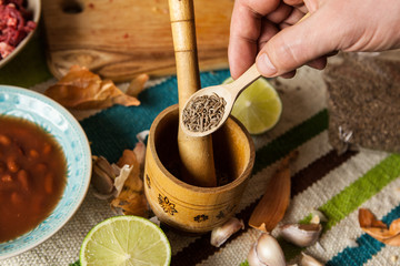 Man's hand holding wooden spoon with cumin seeds above wooden mortar with pestle. Top view, closeup