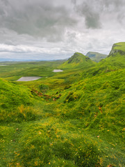 Green hills on the Isle of Skye as seen from Quiraing landslip. Two small lakes at the background with small yellow flowers in front. Overcast weather with grey clouds on a windy summer day.
