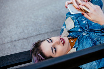 Beautiful urban girl lying on a bench. Looking at smart phone.