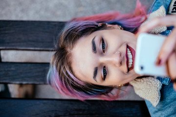 Close-up portrait of a smiling girl with dyed hair using phone while lying on a bench. Top view. - obrazy, fototapety, plakaty