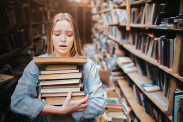 Girl is carrying a lot of books in her hands. It is hard for her to do. She looks hepless and tired. The girl stands in big old library.