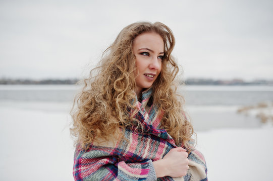 Curly blonde girl in checkered plaid against frozen lake at winter day.