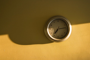 Analogue clock on the yellow wall showing 7:14 time, light from the side, long shadows emphasising...