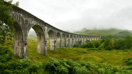 Fototapeta na wymiar High resolution panorama of Glenfinnan railway viaduct (location from Harry Potter movie) shot from side, on overcast day with grey sky and lot of green grass and trees around. Inverness, Scotland