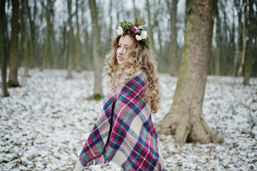 Curly cute blonde girl with wreath in checkered plaid at snowy forest in winter day.