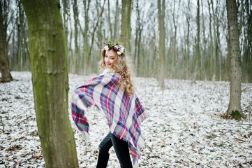 Curly cute blonde girl with wreath in checkered plaid at snowy forest in winter day.