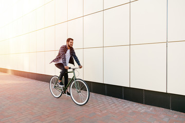 Full length photo of smiling young bearded man riding a bike on the city street. Dressed up in...
