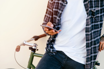 Stylish man chatting while using mobile phone while sitting on bicycle, outdoors. Dressed up in plaid shirt, t-shirt and jeans. Close-up.