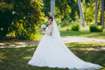 Fototapeta premium Beautiful wedding photosession. The bride in a white lace dress with a long plume, hairpin in hair, veil and bouquet of white and pink flowers in a large green garden on weathery sunny day