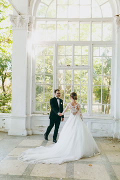Beautiful wedding photosession. Groom in a black suit and young bride in white lace dress with long plume with an exquisite hairstyle in vintage interior on the veranda of an old house with columns