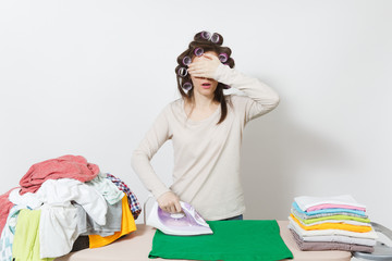 Distressed housewife with curlers on hair in light clothes ironing family clothing on ironing board...