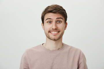 Studio portrait of attractive caucasian guy feeling awkward and confused, not knowing what to say and smiling nervously, standing against gray background, being in strange situation