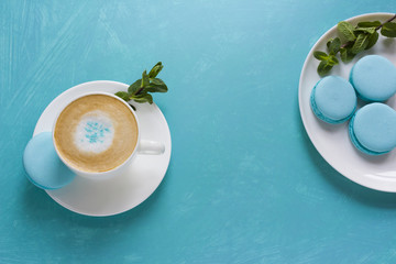 A cup of cappuccino coffee is decorated with mint sugar powder, macaroni cakes on a plate and mint leaves on a blue table is the concept of mint freshness and coffee dessert on a hot day.