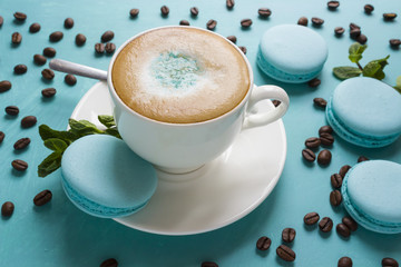 A cup of cappuccino coffee is decorated with mint sugar powder, macaroni cakes, mint leaves and coffee beans on a blue table is a concept of mint freshness and pleasant coolness on a hot day