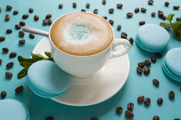 A cup of cappuccino coffee is decorated with mint sugar powder, macaroni cakes, mint leaves and coffee beans on a blue table is a concept of mint freshness and pleasant coolness on a hot day