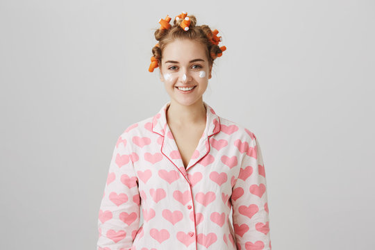 Treat yourself. Portrait of modern cute young woman in hair curlers and pyjamas, standing with facial cream on cheeks and nose, ready to wipe it into skin to make it beautiful, standing over gray wall