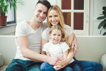 Obraz premium Happy young beautiful smiling parents with a little blond daughter embrace and play sitting at home on the couch. The concept of family