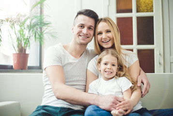 Obraz premium Happy young beautiful smiling parents with a little blond daughter embrace and play sitting at home on the couch. The concept of family