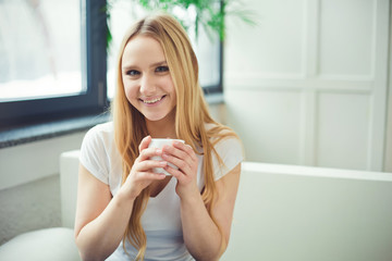 Portrait of a beautiful young blond woman sitting at home on a sofa and drinking hot coffee.