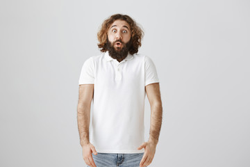 Studio shot of handsome easter man with curly hair and beard standing shocked with dropped jaw and popped eyes, being astonished with kid that jumped from corner and scared him over gray background