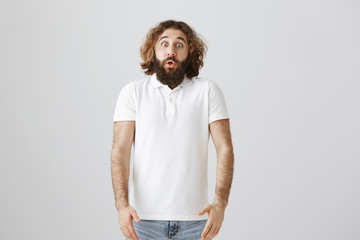 Fototapeta premium Studio shot of handsome easter man with curly hair and beard standing shocked with dropped jaw and popped eyes, being astonished with kid that jumped from corner and scared him over gray background