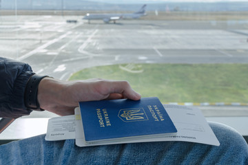 Man’s hand is holding Ukrainian foreign passport with tickets to airplane in background parked...