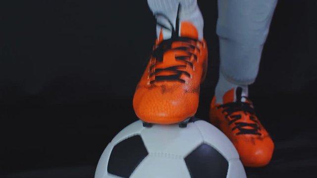 Footballer in boots stop a ball and putting his leg on it, black background