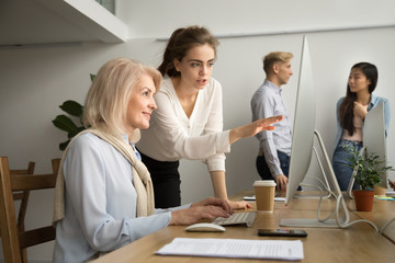 Young businesswoman teaching smiling senior colleague using computer explaining applications work,...