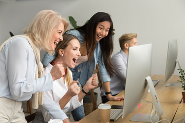 Diverse young and senior female colleagues excited by online win or business achievement, multiracial happy businesswomen celebrating victory looking at computer screen watching cheering winning team