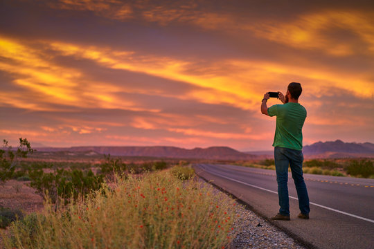 man using smartphone to take photo of colorful sunset beside nevada road in desert