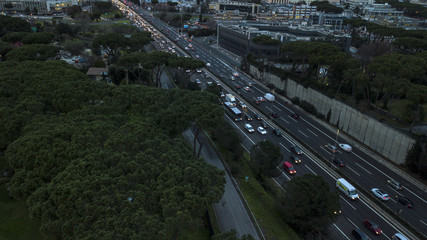 Aerial night view of a highway junction in Rome, Italy. The street crosses an industrial area. On the road, many cars drive at high speed with the headlights on. Around there are grass and trees.