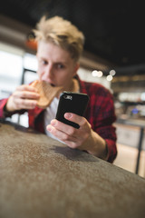 A funny young man uses a smartphone and eats a sandwich in a cozy cafe. Focus on your phone