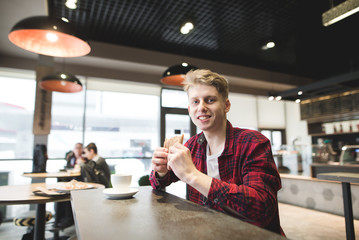 A smiling young man sits in a cafe with a cup of coffee, eats a sandwich and looks at the camera. A positive student dining in a cafe