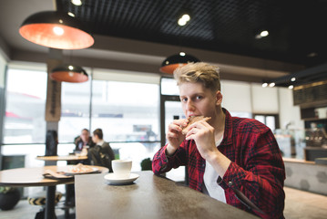 A young man sits in a cafe with a cup of coffee, bites a sandwich and looks at the camera. A positive student dining in a cafe