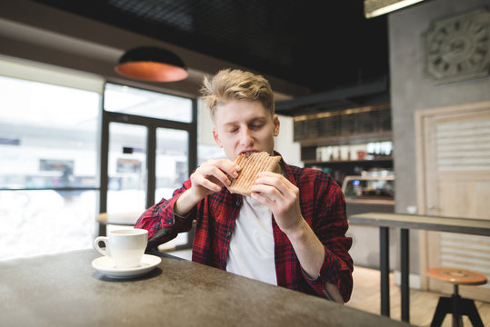 Student with pleasure eats panini sandwich in a cozy cafe. Breakfast with coffee and sandwich. Young man bites sandwich.