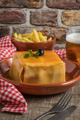 Traditional Portuguese snack food. Francesinha sandwich of bread cheese pork ham sausages with tomato beer sauce and french fries. With a glass of beer and potatoes. On wooden table.