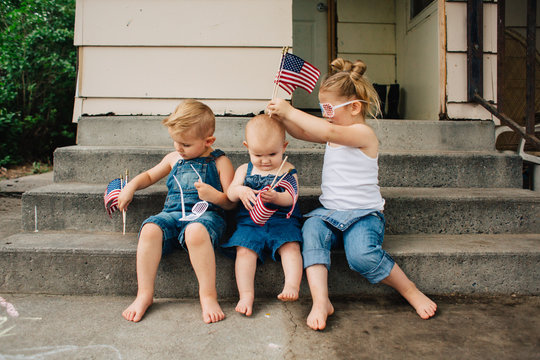 Blonde toddler siblings wearing denim waving American flags outside in the summer.