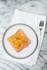 Slice of wholegrain toast with butter, Scandinavian cured or smoked salmon, called Gravad Lax in Sweden, served with mustard and dill sauce on white plate with cutlery on marble plate