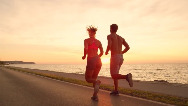 SLOW MOTION, LENS FLARE: Unrecognizable couple jogs down ocean road at sunset. Beautiful evening golden light shines down on athletic man and woman on active date near ocean. Amazing seaside sunrise