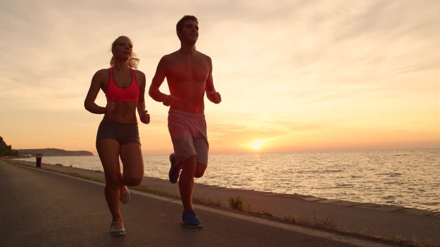 LENS FLARE, COPY SPACE: Cheerful Caucasian couple jogs down scenic ocean road at sunrise. Happy young couple enjoys picturesque evening run along the stunning golden-lit coastline during their holiday