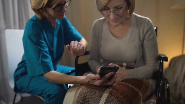 Friendly volunteer teaching woman in wheelchair to use smartphone, nursing home