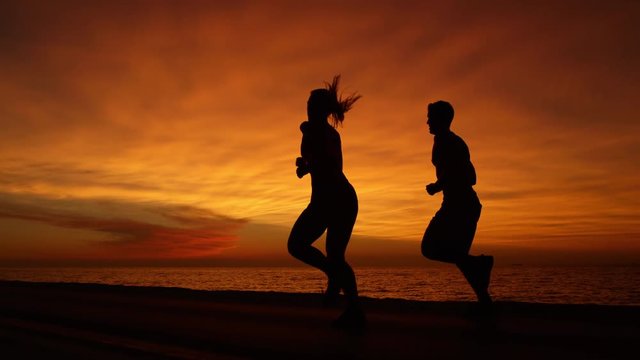 SLOW MOTION, COPY SPACE, SILHOUETTE Young man and woman doing their evening workout in front of tranquil ocean water at gorgeous sunset. Perfect shot of couple jogging along the ocean at orange sunset