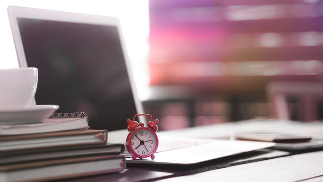 Blurred image, workplace concepts, red color alarm clock with laptop computer and book stack and coffee cup on wooden desk, selective focus for business and education background